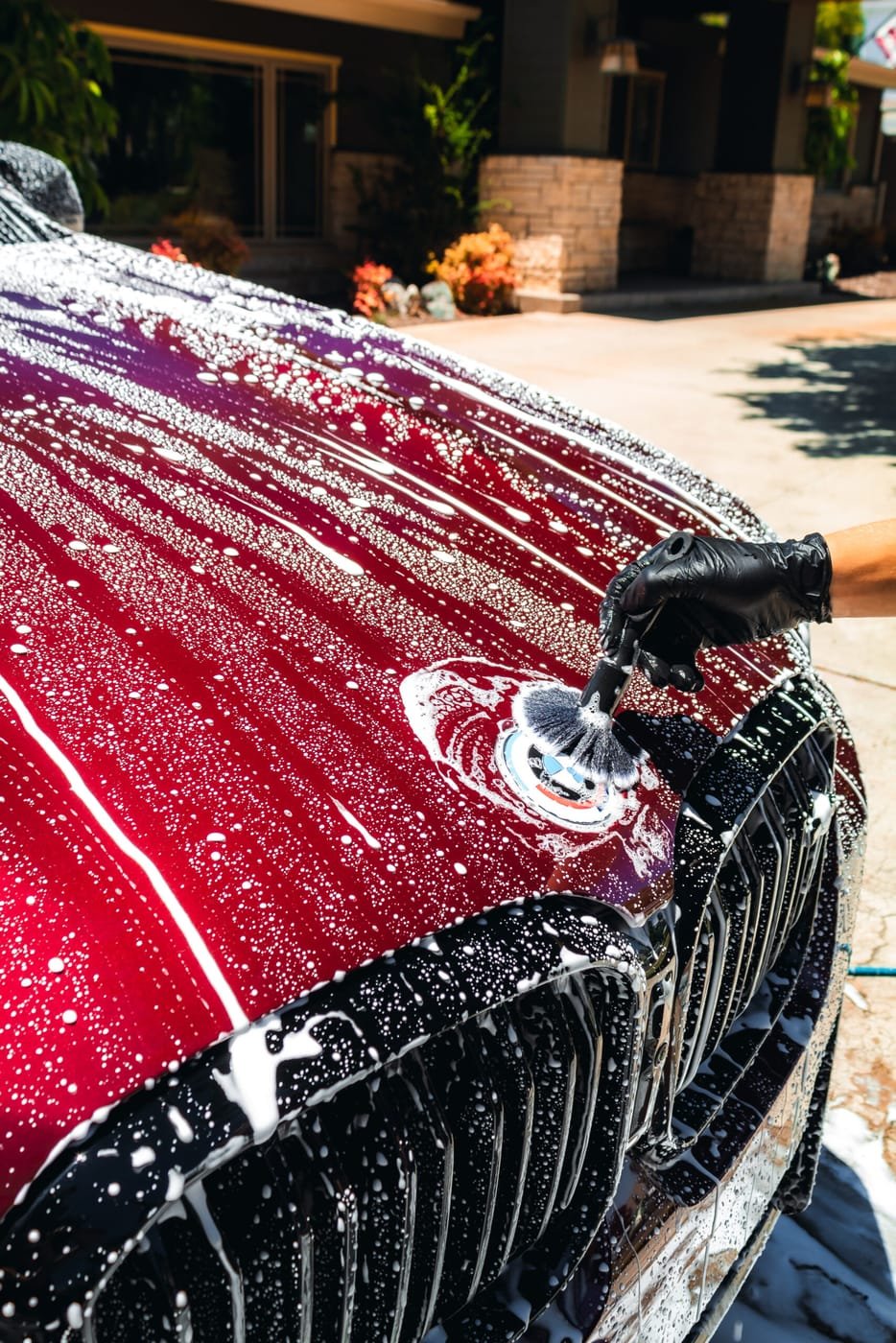 Gloved hand hand-washing a soapy BMW hood and badge with a detailing brush, San Diego driveway
